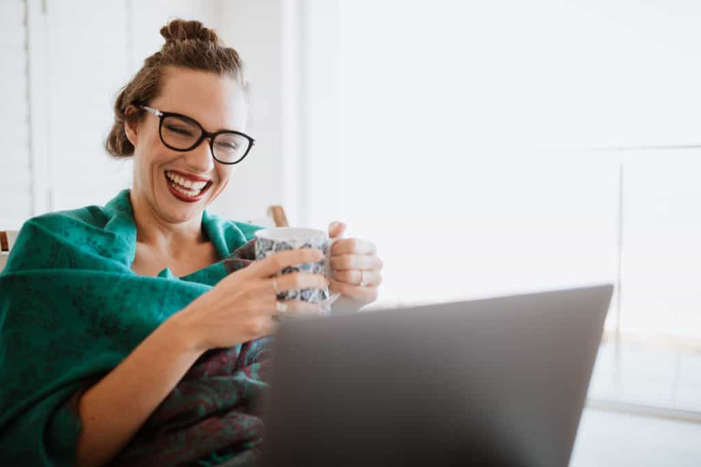 Smiling woman in a green top, holding a coffee mug and looking at her laptop.