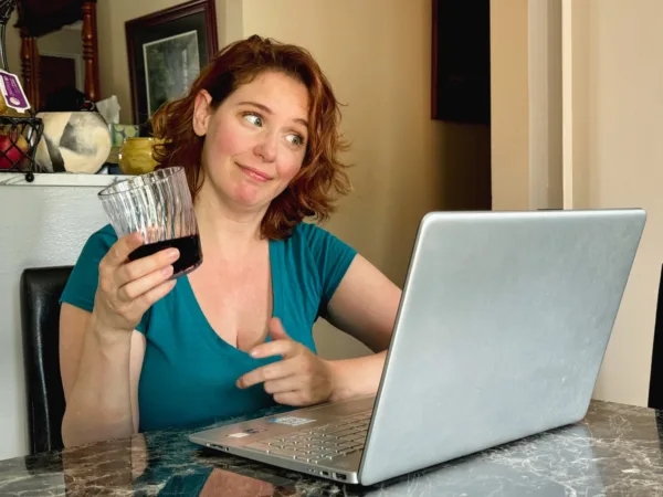 Happy woman with red hair and a green dress holding a glass of wine while playing online poker at a virtual team building party.